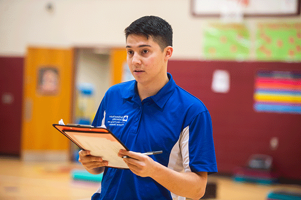 A white male student with dark hair stands in a gymnasium and holds a clipboard and pen. He wears a blue and white Indiana State polo shirt. He looks as if he is asking someone questions. The bottom of a basketball backboard is visible in the background, as is an open door, a maroon-colored wall, and some posters or signage.